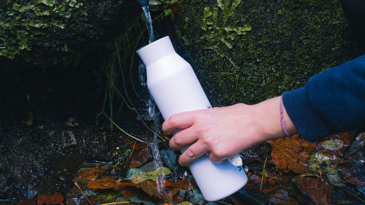 A person filling a white reusable bottle with stream water
